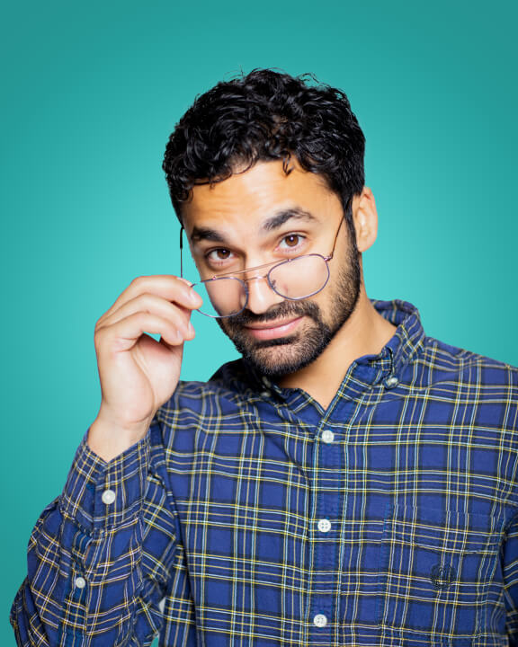A young person in a blue plaid shirt looks earnestly at the camera over the tops of their glasses