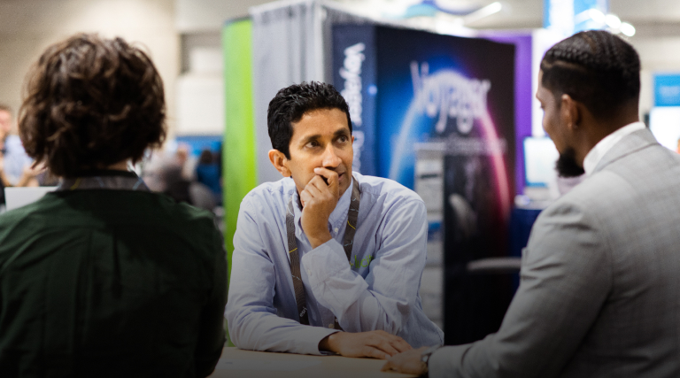 Three professionally-dressed conference attendees grouped around a small table in an expo hall