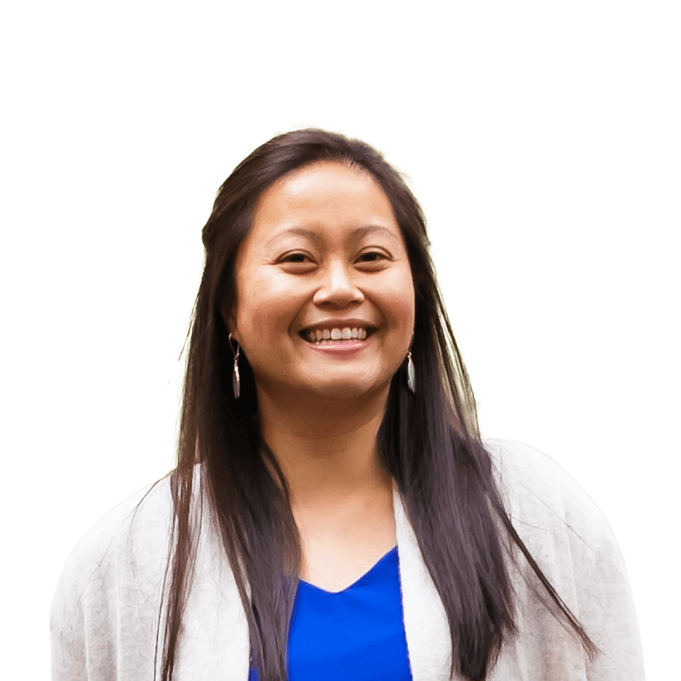 Elizabeth smiling in a blue shirt and white sweater while standing outside beside a grassy field strewn with small trees