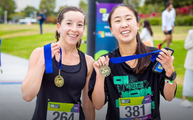 Two smiling people standing outdoors in a field wearing racing bibs and holding medals 