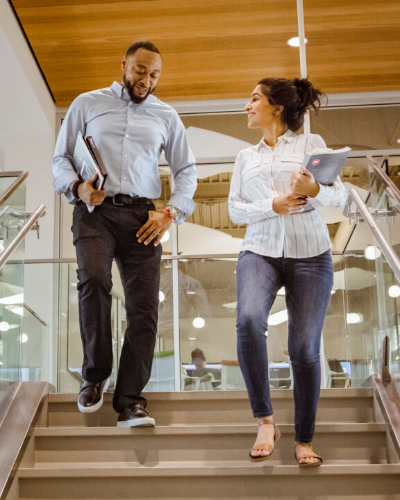 Two smiling people chatting as they descend a pale wood stairwell in a brightly lit modern office building