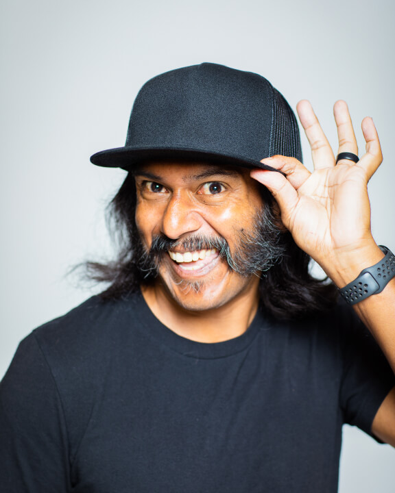 A mustached person in a black tee shirt and baseball cap smiling playfully at the camera