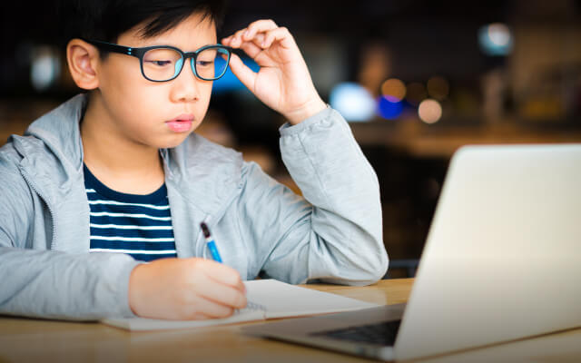 A child in a gray jacket writes with paper and pen while studying at a laptop