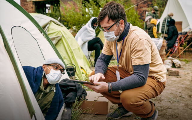 A person holding a clipboard crouches beside a row of tents in a green field as another person leans out of a tent to speak to him