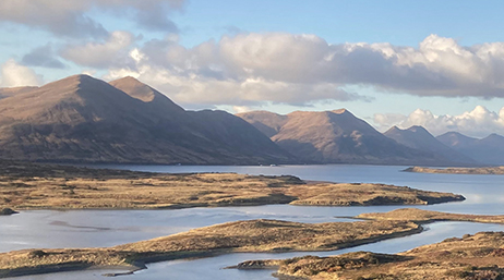 Brown marshlands surrounded by a large body of water at the foot of a range of brown mountains below a partly cloudy sky