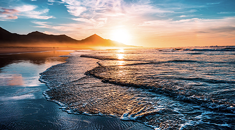 Shallow waves lapping on a shore with mountains in the distance and the sun on the horizon