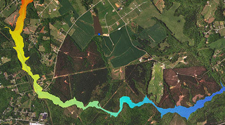 An aerial view of a green rural area with a river winding through the middle, which is shaded in rainbow colors