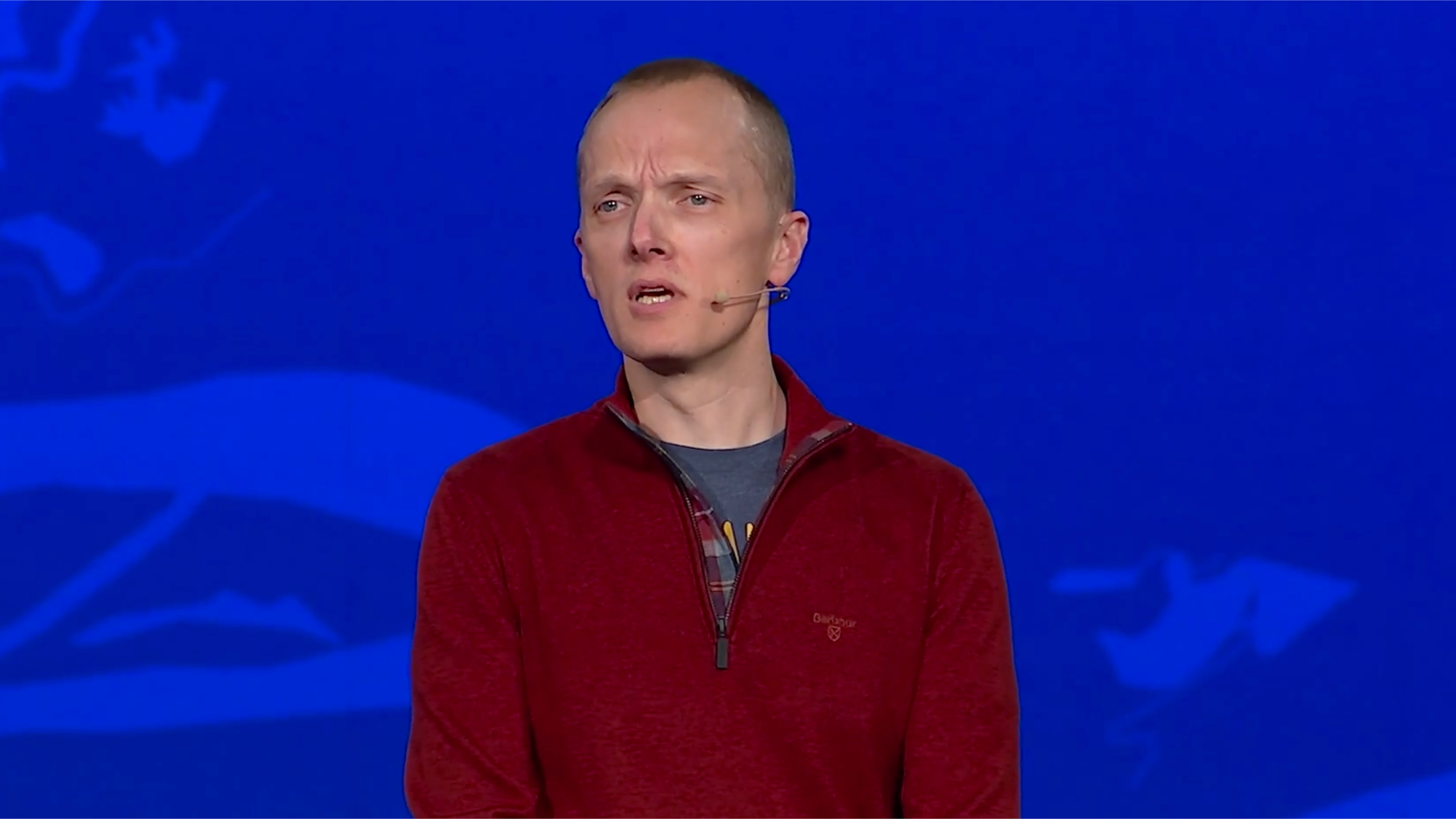 A man in a red sweater speaking in front of a blue background