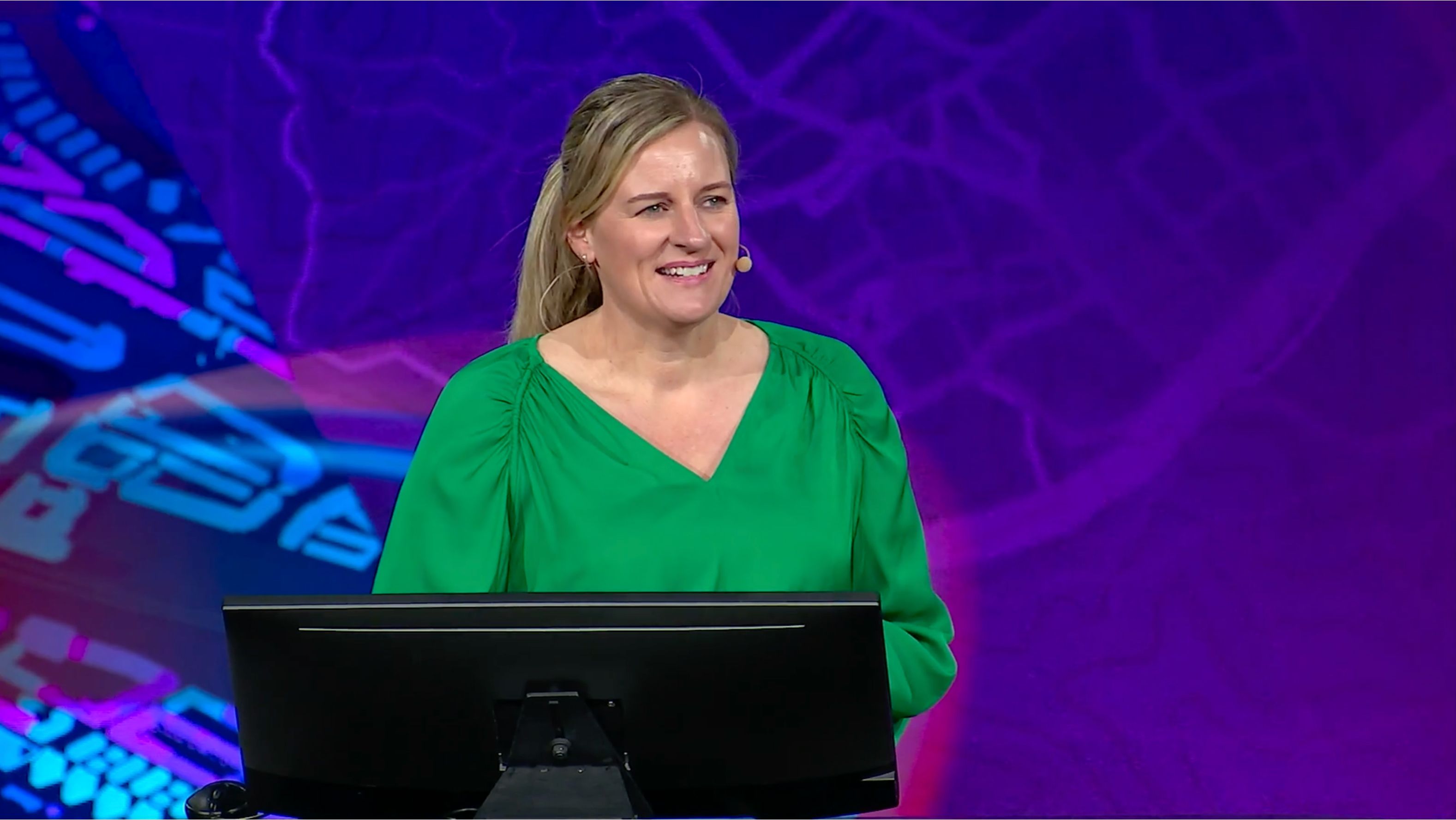 A woman in a green shirt speaks into a microphone in front of a purple background 