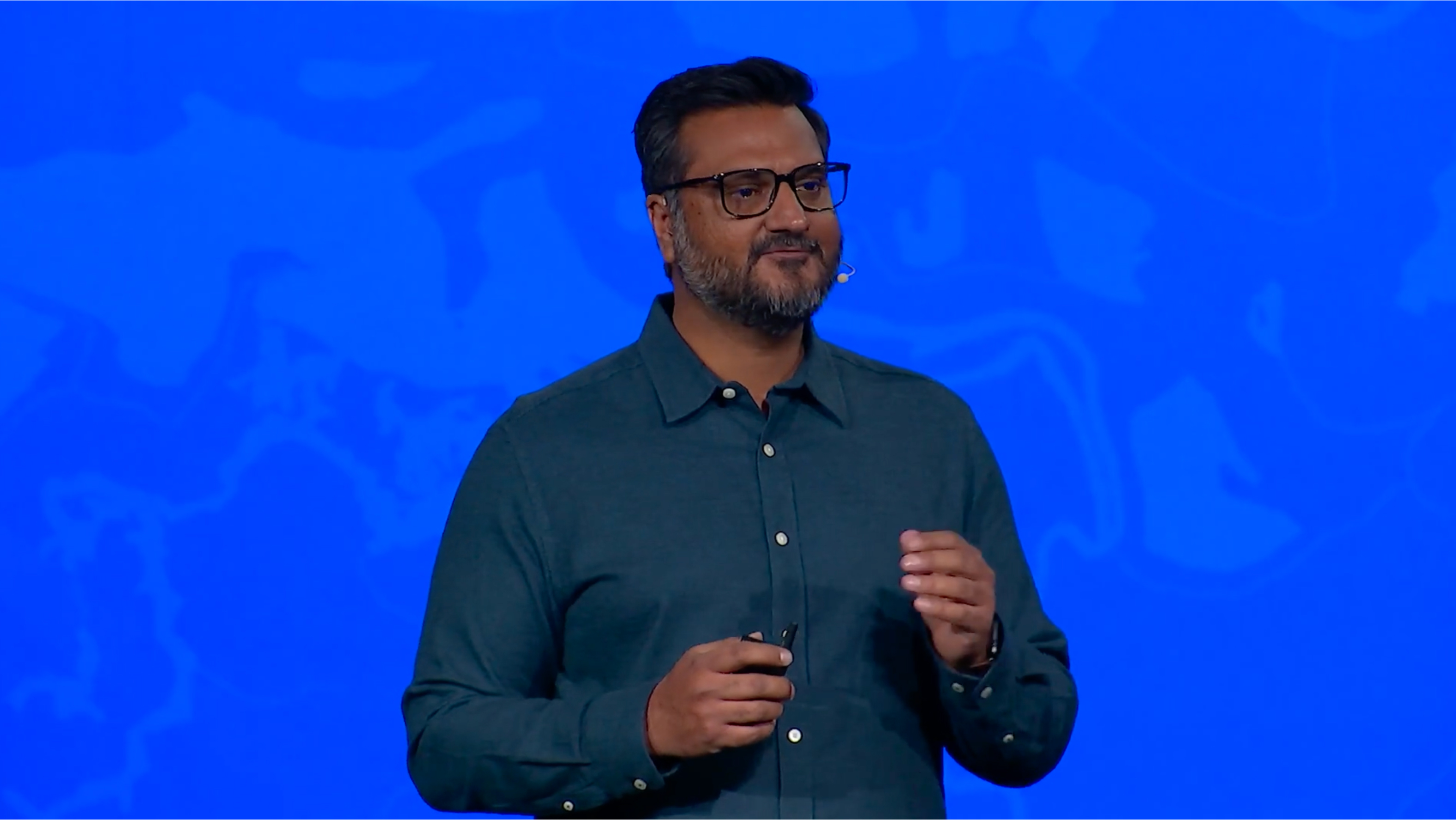 A man in a dark blue shirt and glasses stands in front of a blue screen and talks