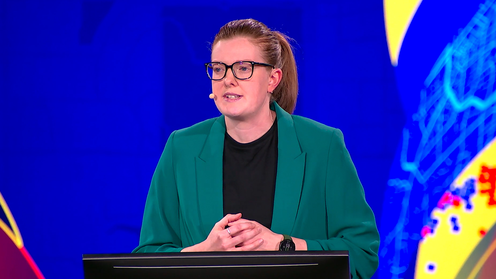A woman in a green blazer and glasses speaks at a lectern, against a blue, yellow, and red background