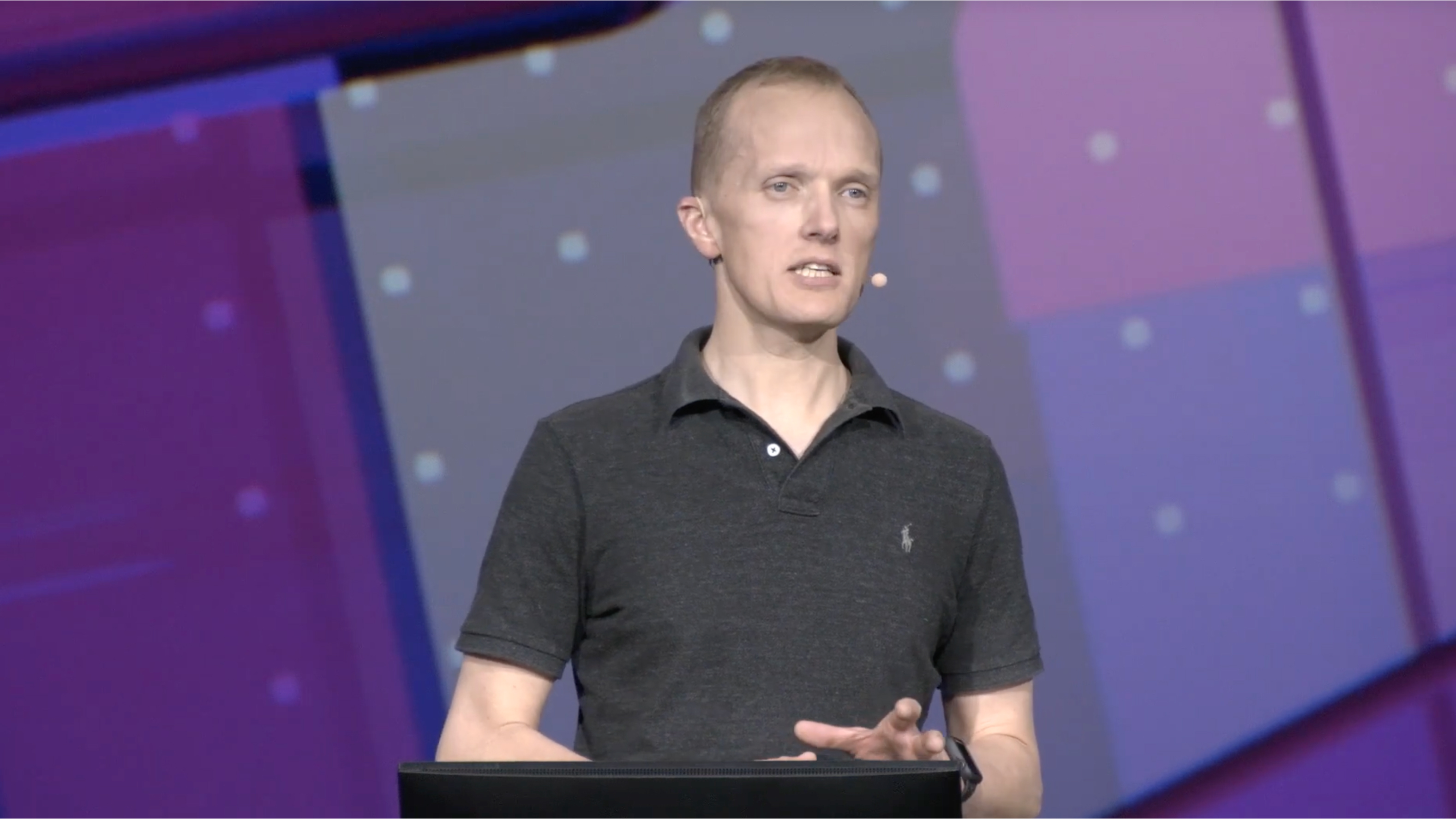 A man in the gray polo shirt is speaking into a microphone at a conference