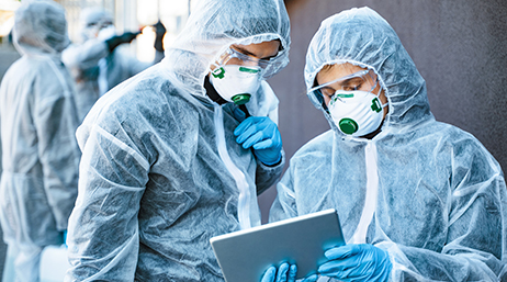 Two people in protective suits with masks and glove talk with others similarly dressed behind them