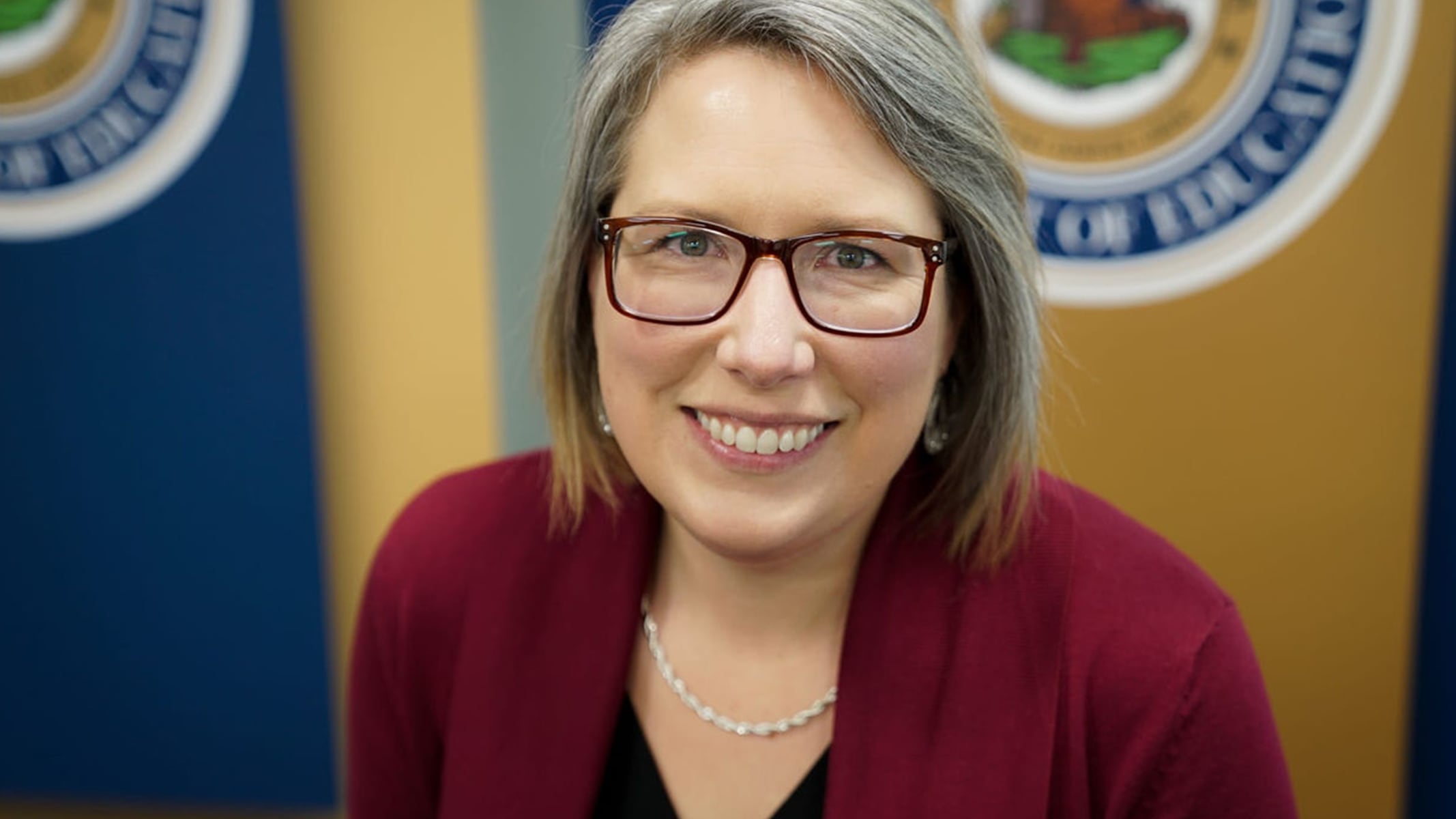 Dr. Klose smiling in a burgundy sweater with a West Virginia Department of Education official seal in the background