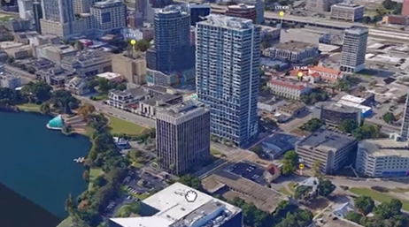 A 3D digital mesh model of an area with multistory buildings near a manmade lake overlaid with a play button