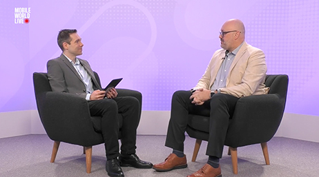 Two people sitting across from each other in armchairs in front of a lavender backdrop overlaid with a play button