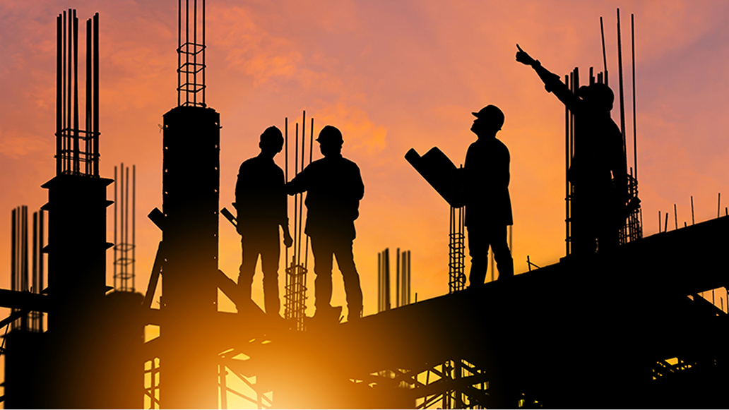 Four people wearing safety helmets and holding tools and blueprints stand silhouetted on the girders of a construction site against a gold and pink sunrise sky